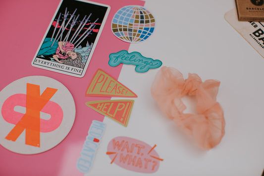 Collection of playful stickers including a tarot card, globe, speech bubbles with text, and decorative designs on a pink and white surface with a peach scrunchie.