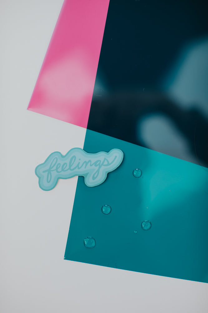 A turquoise 'feelings' sticker placed against a geometric background of pink, teal, and white sections with water droplets on the teal surface"