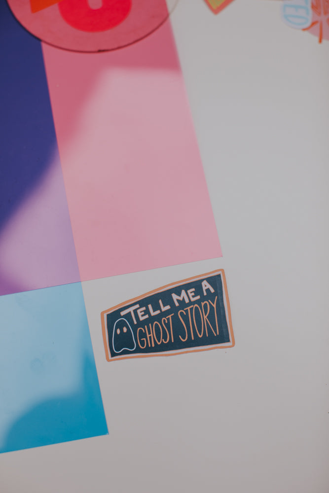 The same 'TELL ME A GHOST STORY' sticker with ghost icon shown against a soft, dreamy background of pink, blue, and white geometric shapes