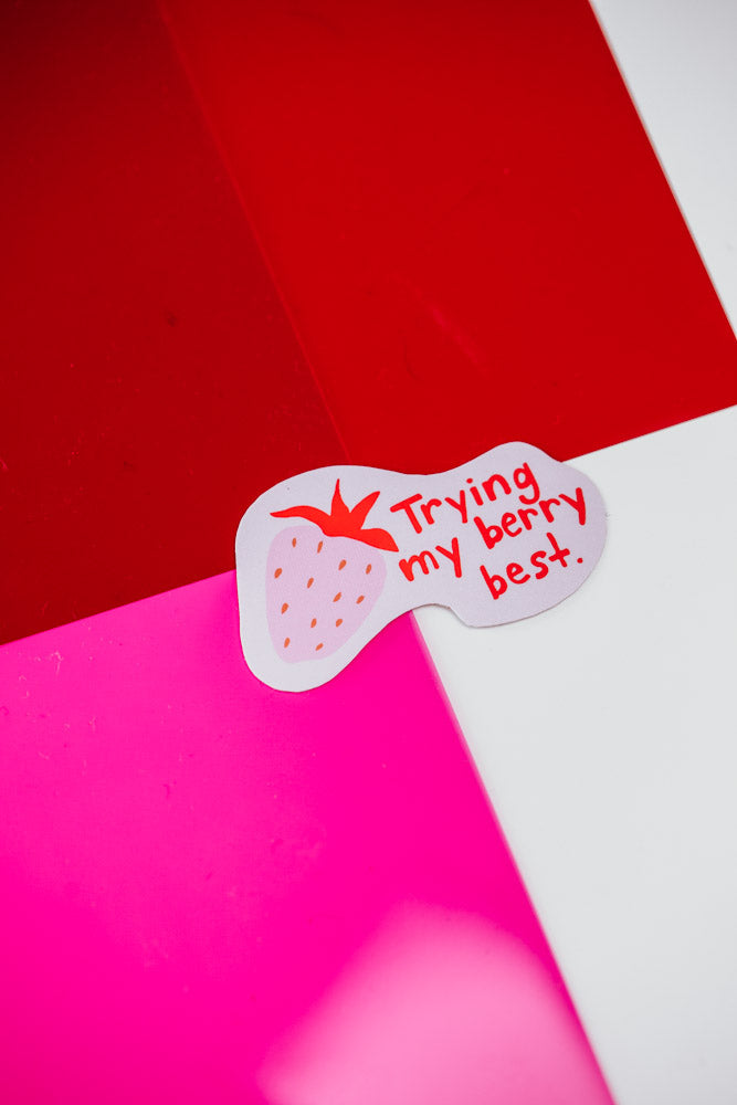 Sticker with a strawberry illustration and text reading "Trying my berry best" placed on red and pink color blocks.