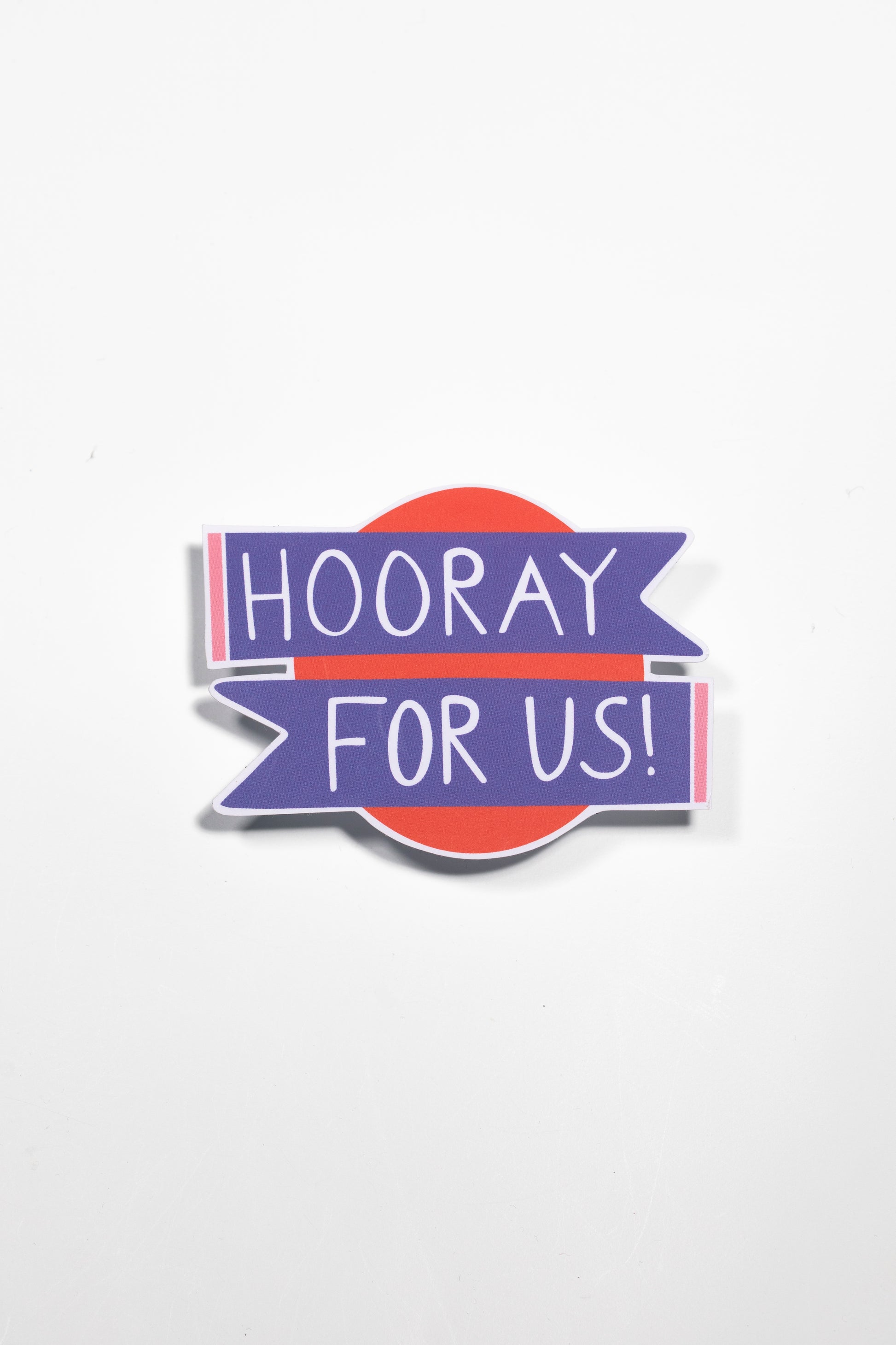 The image shows a sticker with the text "HOORAY FOR US!" written in large, bold purple letters. The text is placed on a purple and orange background with a retro, vibrant aesthetic.