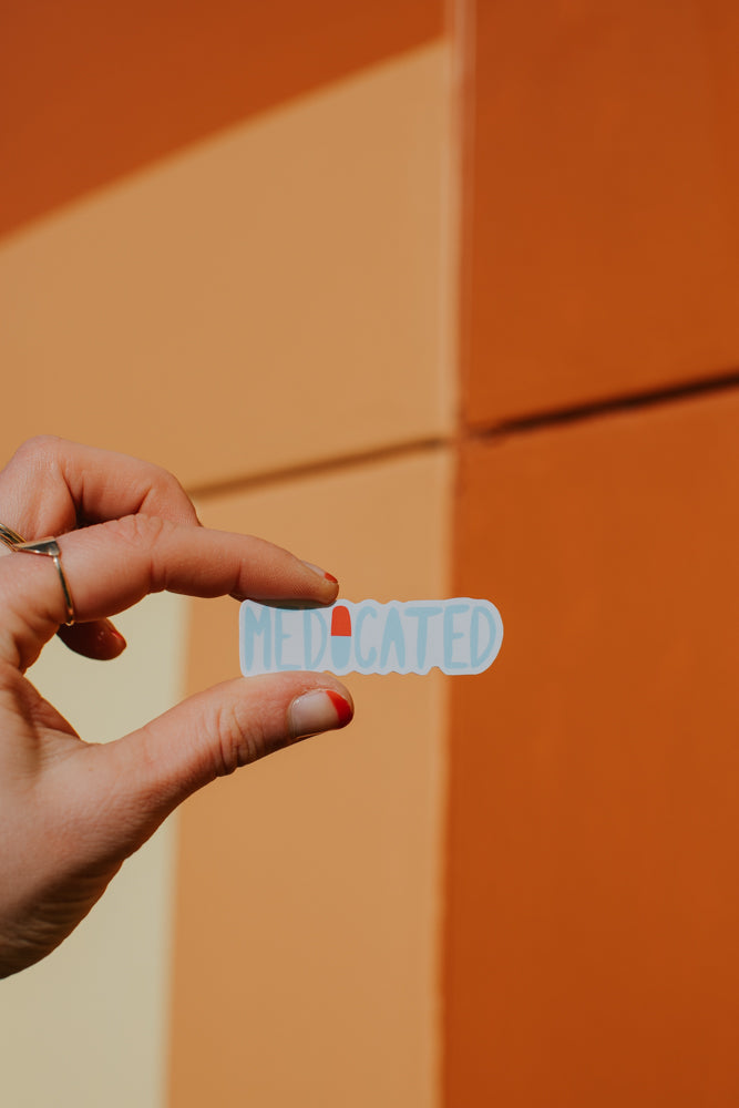 A close-up image showing a hand holding a white sticker or container with the word "MEDICATED" printed in blue and red lettering, against a vibrant orange background.